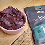 A bowl of Made For Drink's Biltong on a wooden table next to a 30g unopened pack in partial view