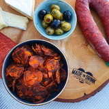 A bowl of Made For Drink's Chorizo Thins on a charcuterie board next to a whole sausage of chorizo, a bowl of green and black olives, some slices of brie and salami