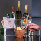 A hand reaching for a bowl of Made For Drink's Dorset Sea Salt Crisps in from of a gin and tonic garnished with dried orange pieces, a box of unopened 40g packs of crisps and a silver ice bucket filled with 3 bottles of champagne.