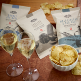 150g packs of Made For Drink's English Truffle, Malt Vinegar & Sea Salt and Dorset Sea Salt flavoured English Heritage potato crisps., lying on a wooden table with a bowl of crisp and 2 glasses of champagne.