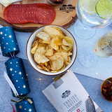 A bowl of English Truffle crisps on a festive set table next to a charcuterie board with salami on, a gin and tonic and glass of champagne.