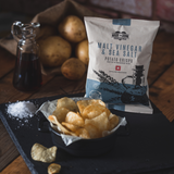 A pack of Made for Drink Malt Vinegar and Sea Sal crisps standing on a slate board with a bowl of crisps in the foreground next to a pile of sea salt. In the background is a small jug of malt vinegar and fresh potatoes in a brown hessian sack