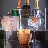 A bowl of Made For Drink's Dorset Sea Salt Crisps standing in from of a gin and tonic garnished with dried orange pieces, a box of unopened 40g packs of crisps and a silver ice bucket filled with 3 bottles of champagne.