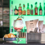 A silver box holding 3 40g packs of Made For Drink's award-winning English Truffle flavoured English Heritage potato crisps, next to a gin and tonic served with berries and garnish and a bowl of crisps on top of a bar with drinks bottles in the background.