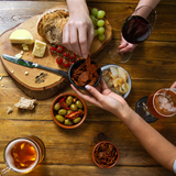 Close up of a spread of snacks on wooden table: a wooden board with cheese, bread, grapes and tomatoes on the vine, a bowl of olives and sundried tomatoes, bowls of chorizo thins and a bowl of crisps, alongside glasses of beer, red wine and hands sharing food.