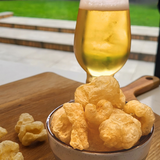 A bowl of Made for Drink's Pork Chicharrones on a wooden serving board with a pint of beer in the background.