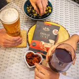 A 3og packet of Chorizo Thins in the middles of a white rattan table with a small heart shaped bowl filled with thins and a large bowl of mixed olives and feta cheese.